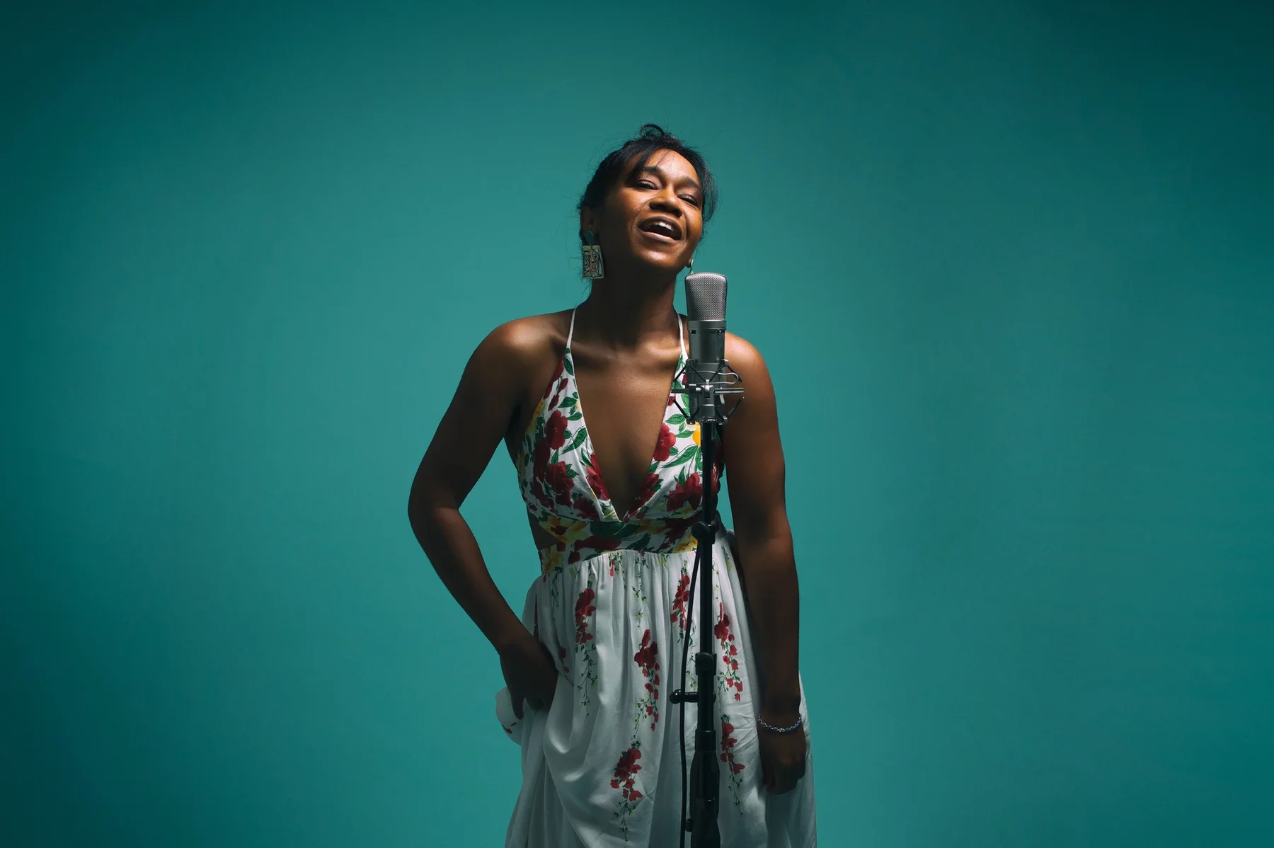 Magela Herrera singing joyfully at a studio microphone in a white floral dress, against a teal backdrop