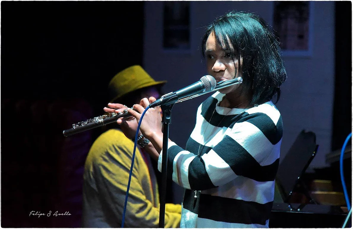 Magela Herrera playing flute into a microphone on stage, wearing a black and white striped top