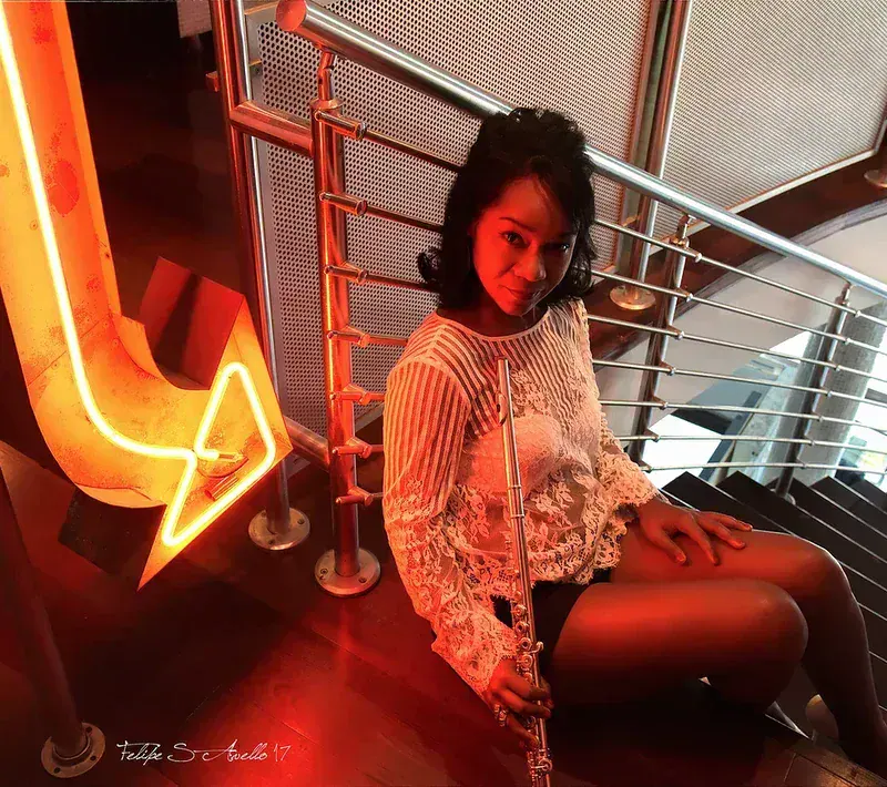Magela Herrera seated on a staircase holding her flute, beside an orange neon arrow