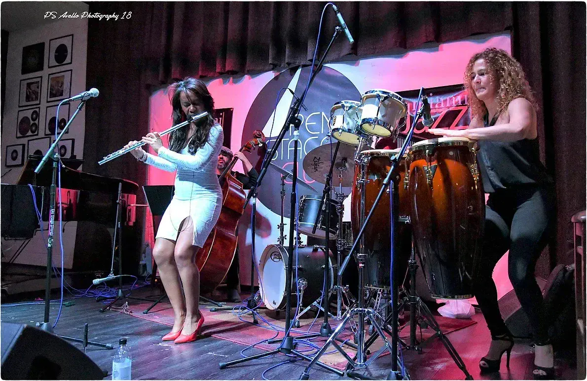 Magela Herrera playing flute on stage beside a percussionist on congas, with bass and drums behind