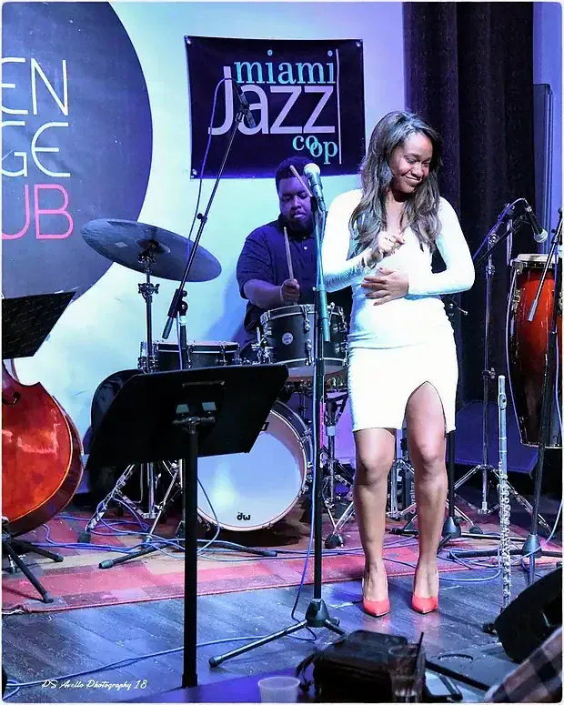 Magela Herrera smiling on stage in a white dress with a drummer behind her