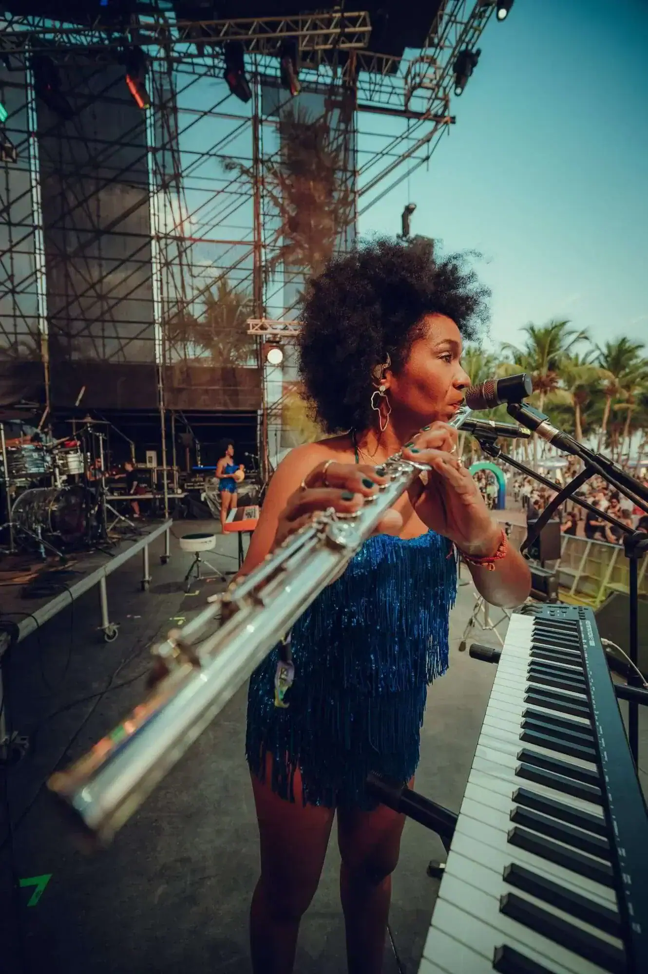 Magela Herrera playing flute on an open-air stage with palm trees, beside a keyboard and facing the audience